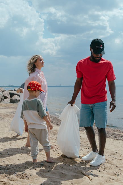 A man wearing a black cap, red t-shirt, and light blue denim shorts is standing on a sandy beach, holding a white rubbish bag in one hand. Beside him, a young boy in a red cap, grey and white outfit, 