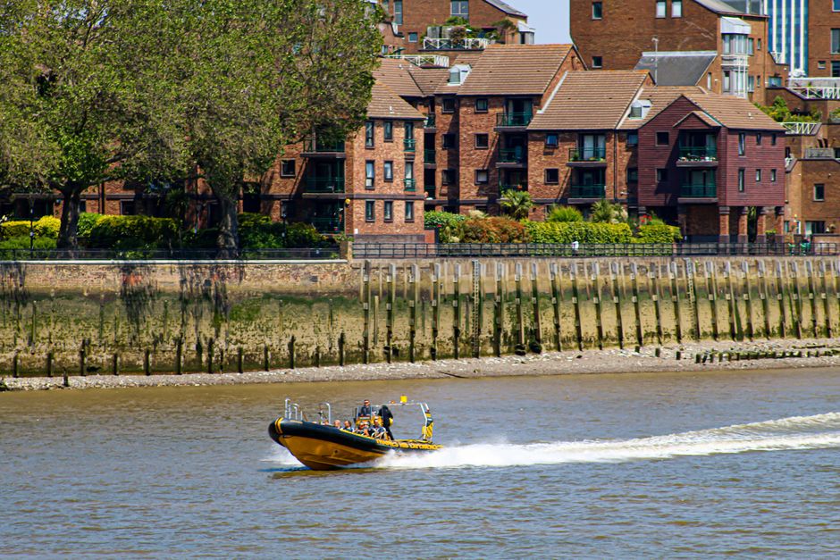 A yellow and black rigid inflatable boat speeds across a river, creating a trail of white foam behind it. The boat is occupied by several people wearing life jackets, and it appears to be part of a wa