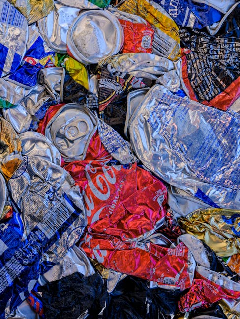 Close-up view of a pile of crushed aluminium cans, primarily from soft drink brands such as Coca-Cola, with some cans featuring distinctive red, blue, gold, and silver colours. The cans are tightly co